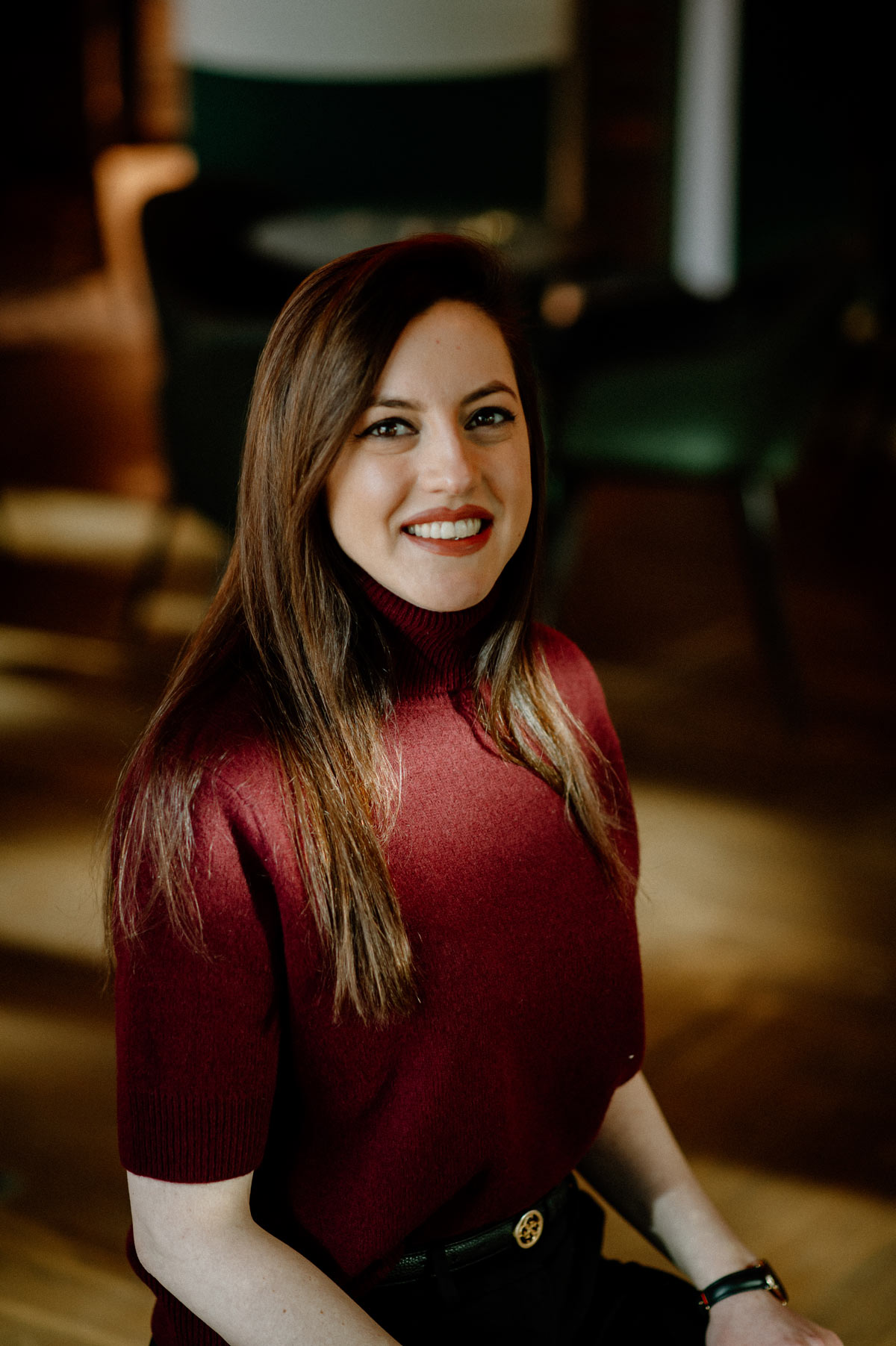 Junge Frau mit langen braunen Haaren lacht freundlich im bordeauxfarbenen Pullover